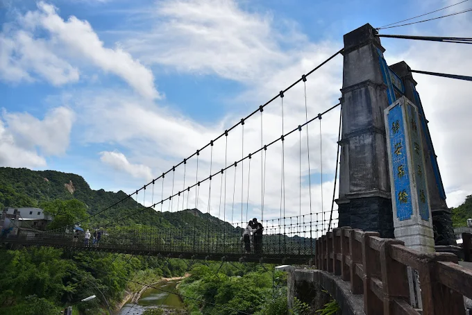 遊客走在新北平溪靜安吊橋上，背後是綠意山巒與藍天白雲
