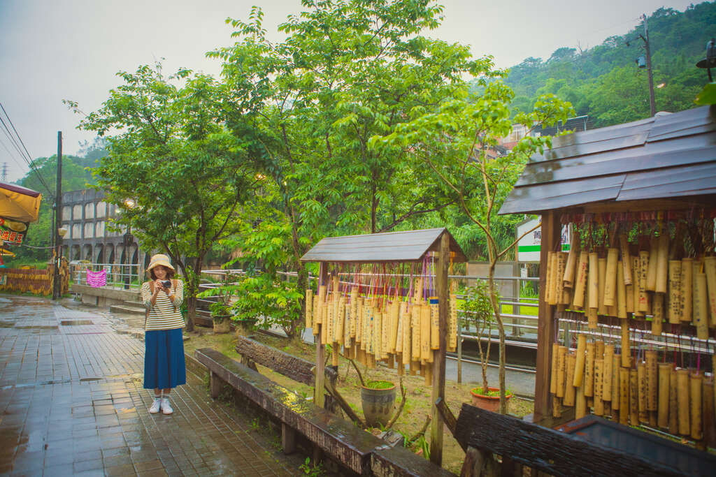 菁桐老街一名旅客站在祈福竹筒牆前拍照，街道濕潤安靜，綠意盎然

