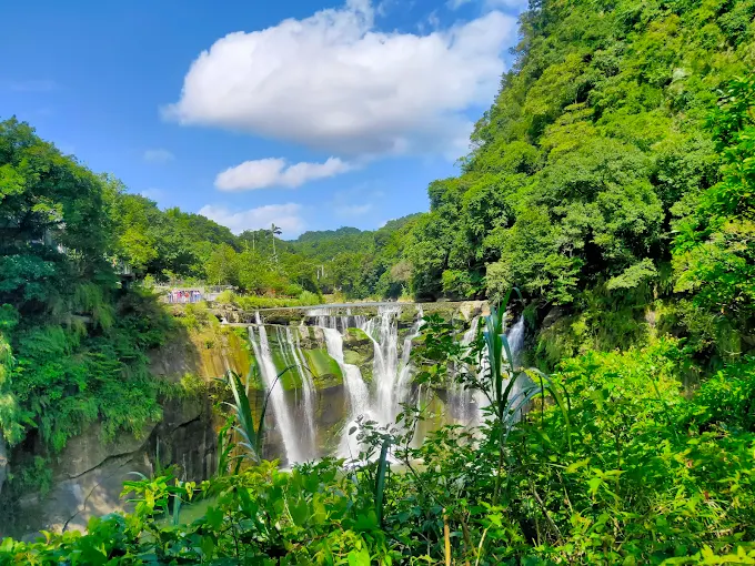 十分瀑布在藍天白雲映襯下流瀉而下，前景為翠綠樹葉
