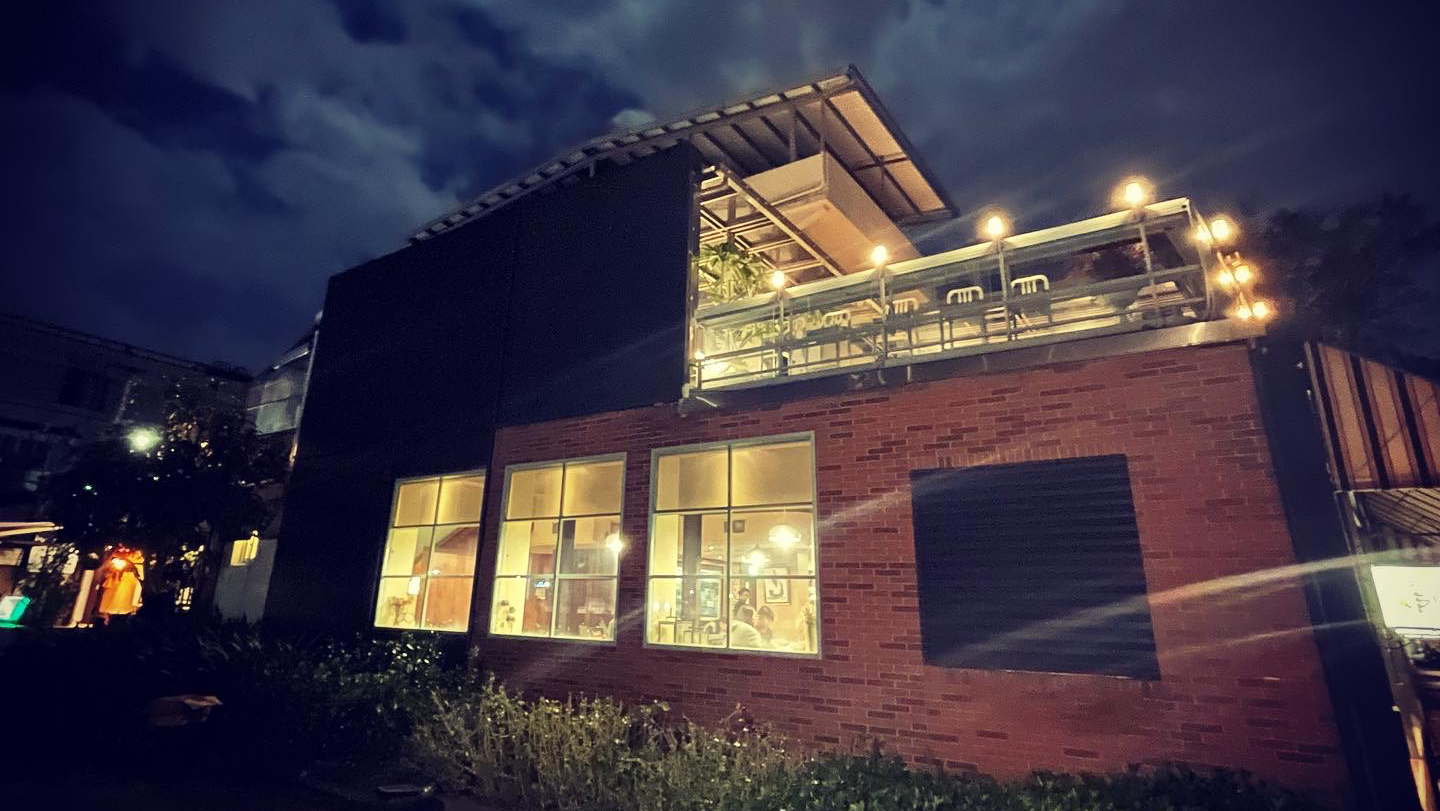 夜晚燈光溫暖的磚造建築，二樓有露天陽台座位區，周圍有綠植