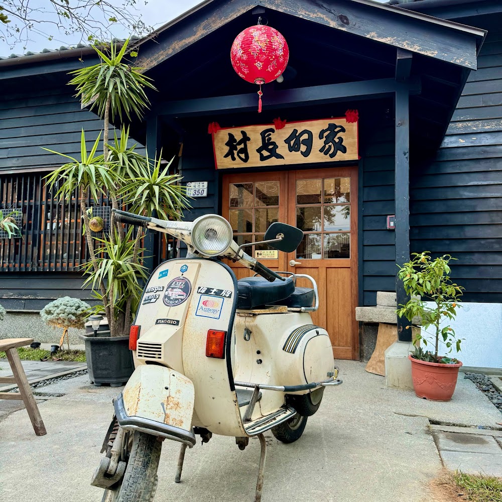 黑色木板老屋外觀，掛著村長的家招牌與紅燈籠，門前停著白色復古機車