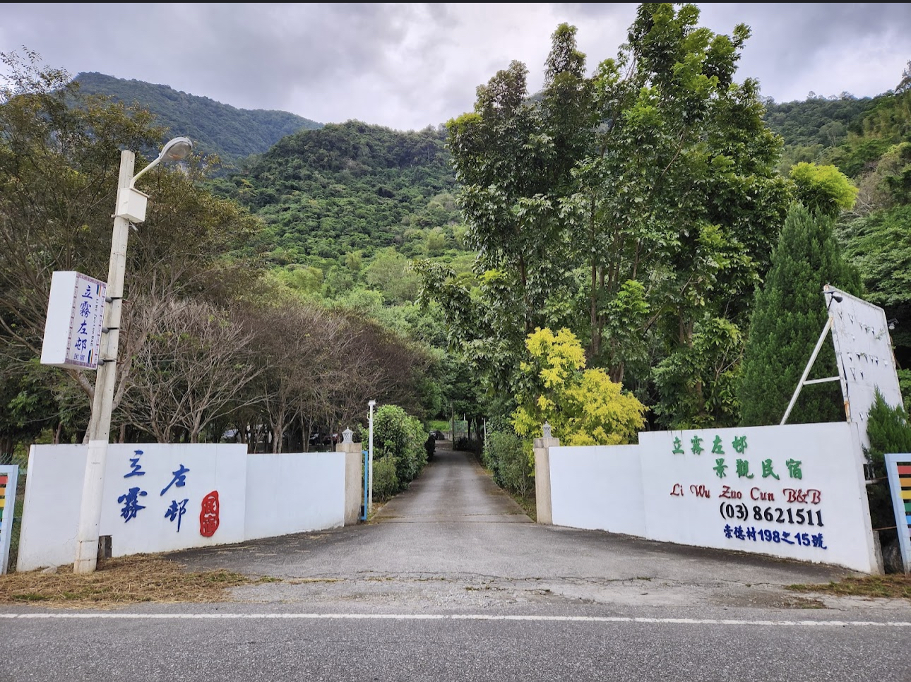 民宿入口四周環繞著茂密綠樹與青山，入口處有白色圍牆與招牌