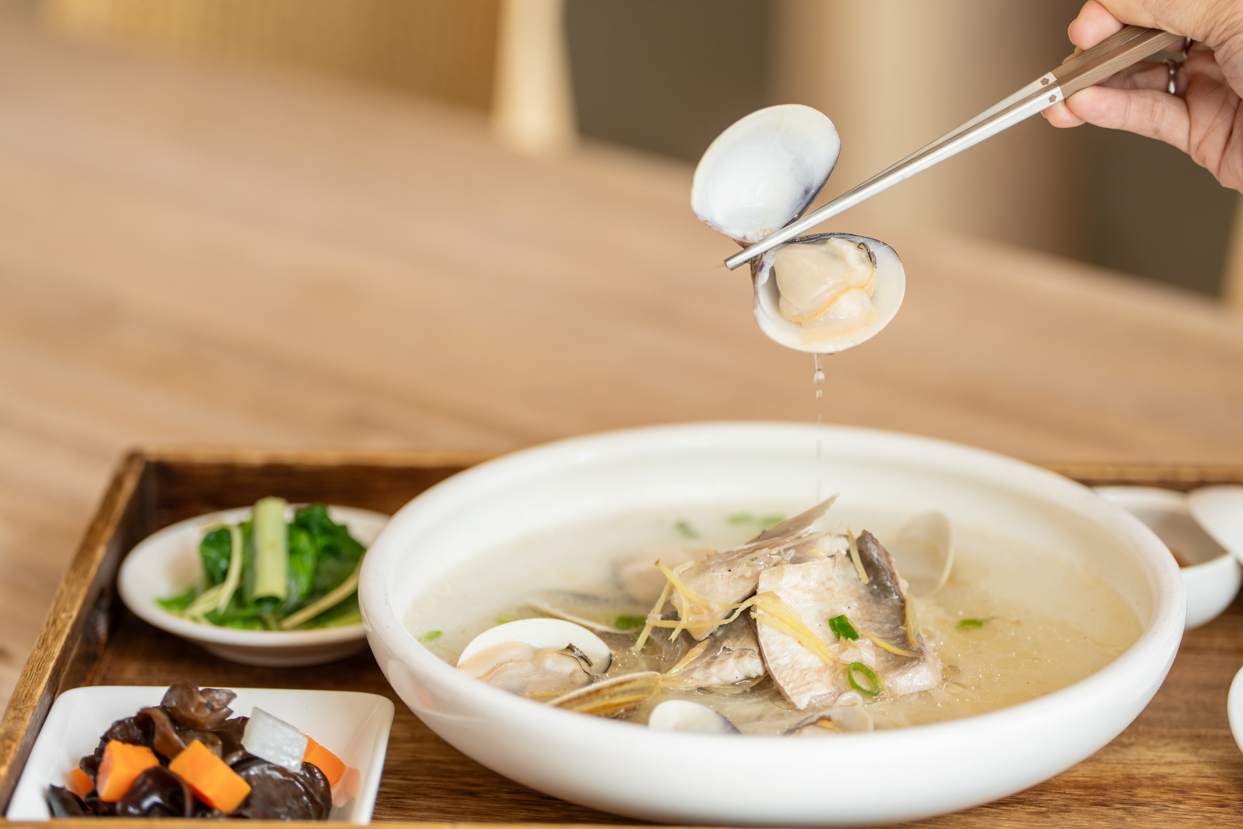 這張圖片主要是一碗鮮美的魚頭與蛤蜊湯，湯裡有魚肉、蛤蜊及蔥花，旁邊還有小菜如木耳、胡蘿蔔與青菜，呈現豐富的海鮮與蔬食搭配。