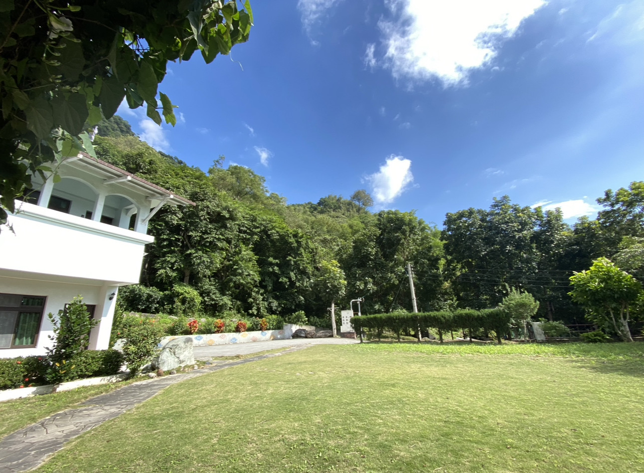 白色建築物旁有大片綠草地，四周是樹林與藍天白雲