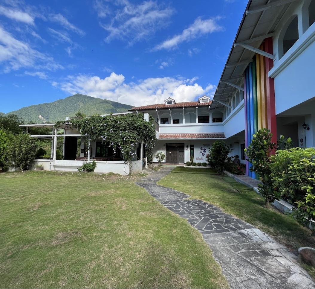 白色建築外牆有彩虹色裝飾，前方是綠色草地和小徑，背景有山景和藍天白雲