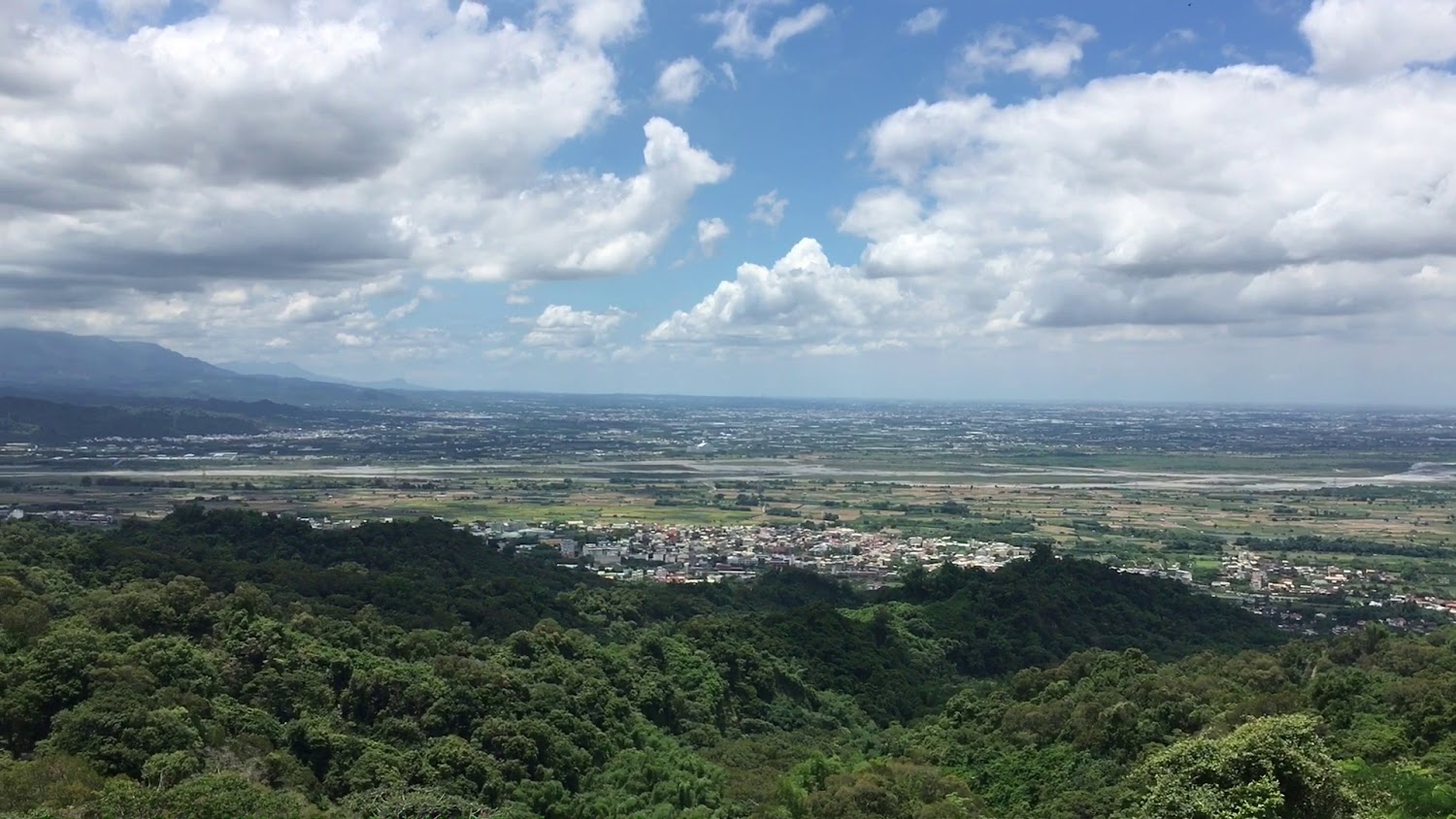 一片廣闊的田野和農村聚落，前景有大片綠色森林，遠方可見平原和分布的房舍，上方是藍天白雲，非常壯闊自然。