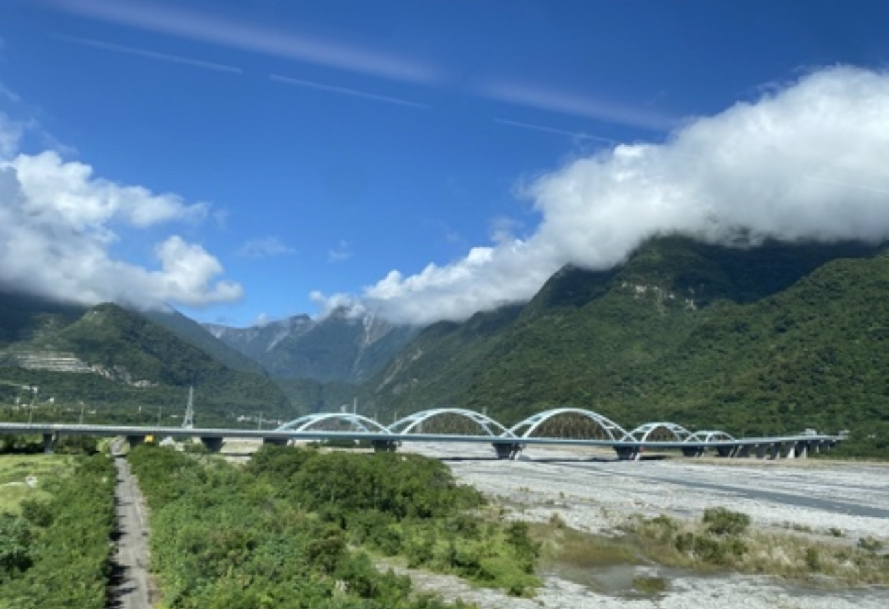 藍色拱橋橫跨在河床上，背景是青翠山脈與藍天白雲
