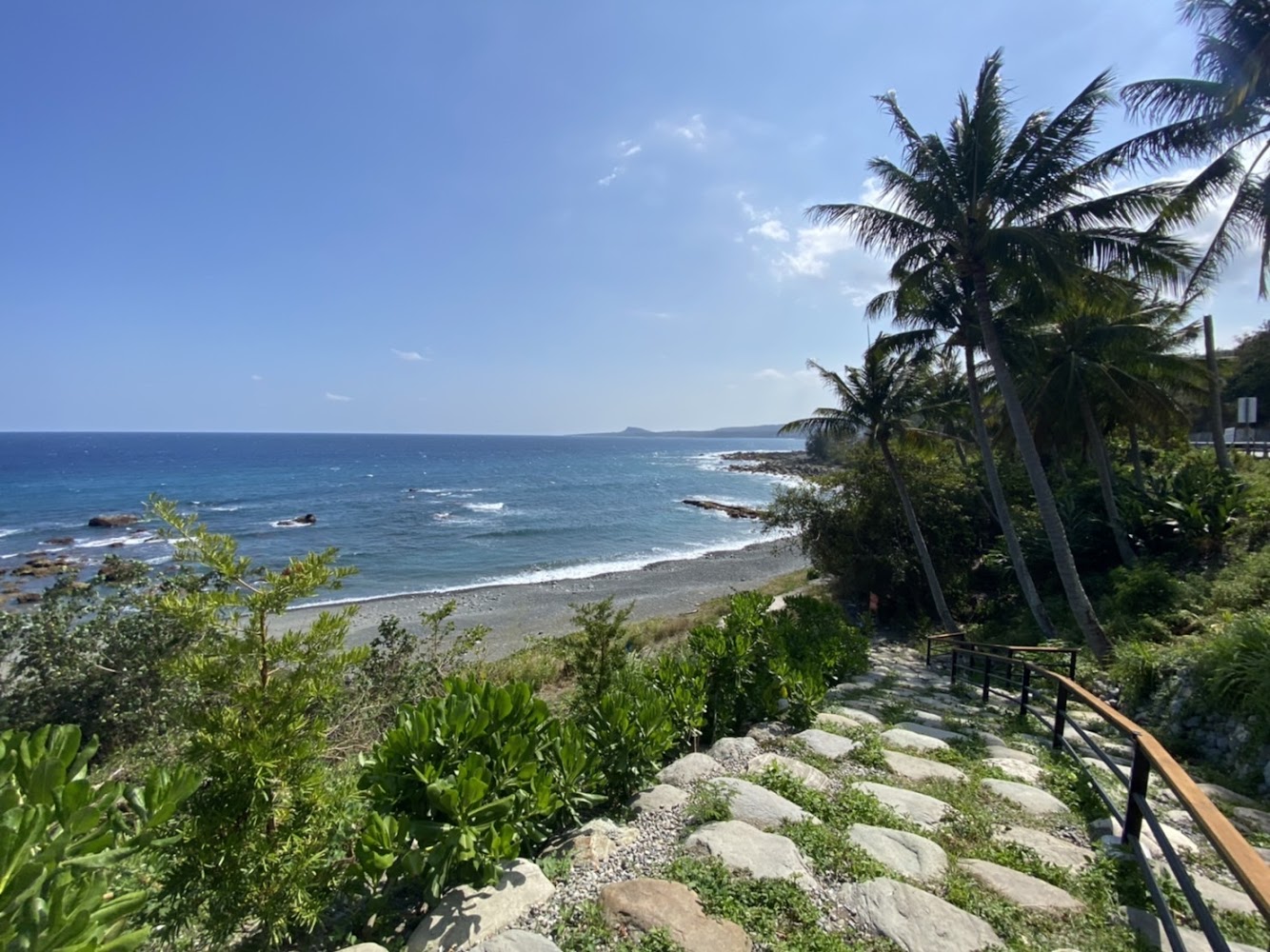 蔚藍的海洋、沙灘和礁石，右側有椰子樹及綠色植被，前景是一條石階步道通向海邊