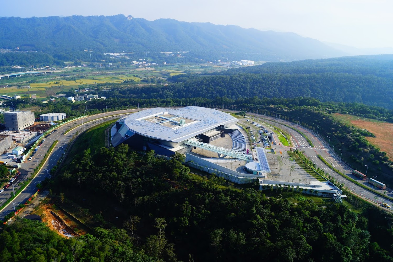 這張圖片主要呈現一座現代化的建築，建築周圍有大片綠林和山脈，遠方可見農田和城市景觀，環境清幽，融合自然與人文設計。
