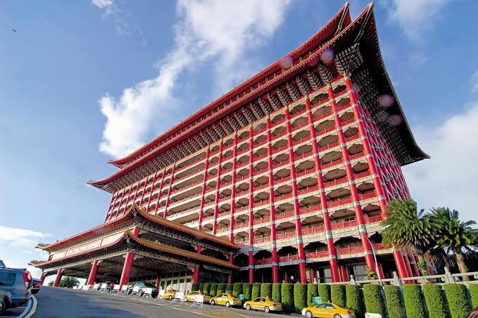 圓山大飯店正面外觀，仰角拍攝宮殿式紅色建築，前有計程車及修剪整齊綠籬，藍天白雲