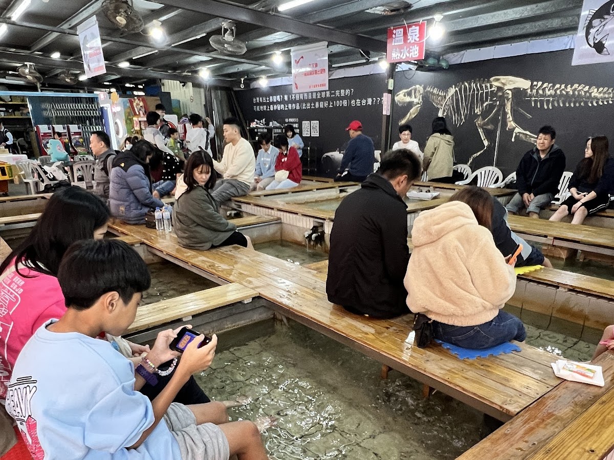 湯圍溝溫泉魚SPA體驗，室內木製長凳，遊客坐著泡腳，黑色壁面有恐龍骨架壁畫