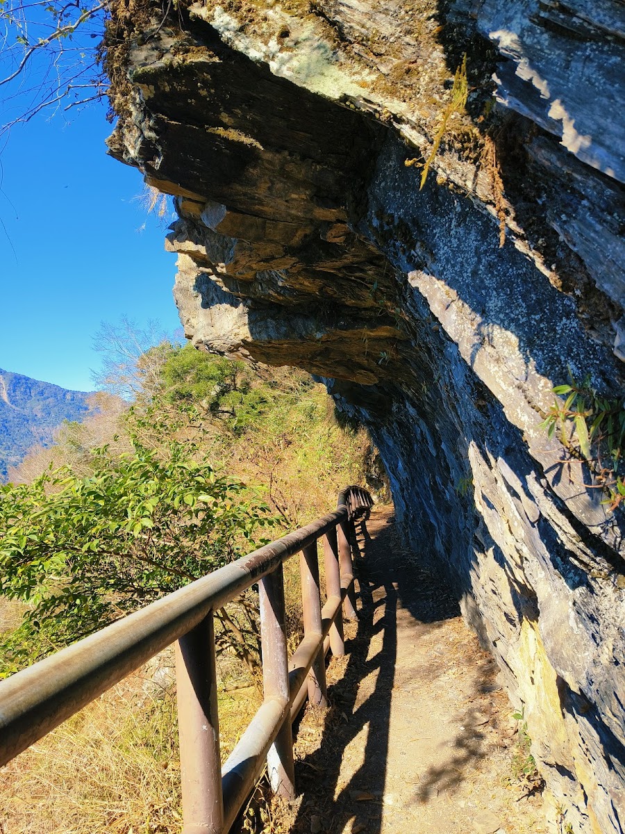 山壁懸崖旁的步道，右側緊貼岩石，左側有鐵製欄杆，遠方是藍天山林