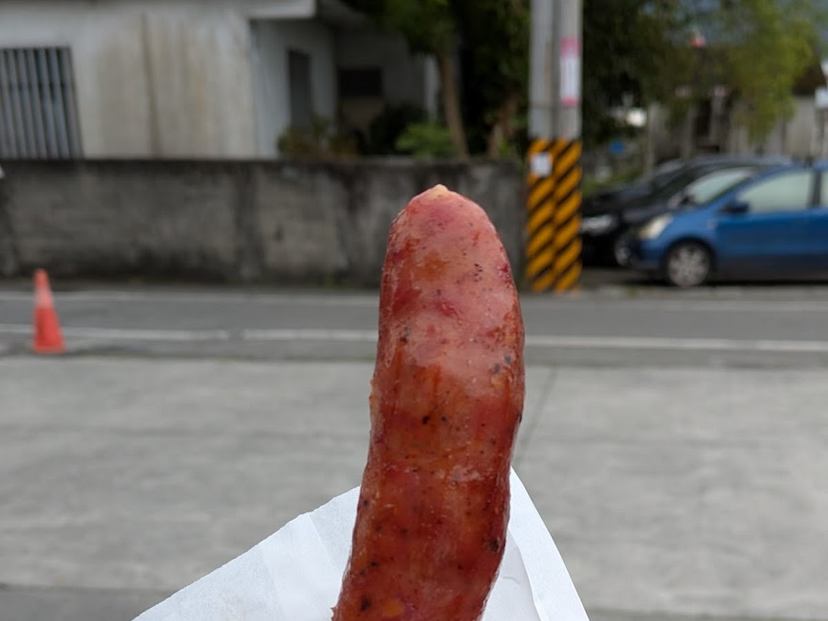 手持馬告香腸竹籤，以白色包裝紙托著，停車場取餐情境