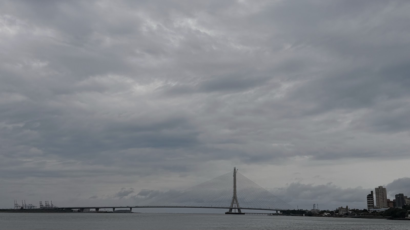淡水河全景，陰天，斜拉橋橫跨河面，左側有港口起重機，新北市景觀