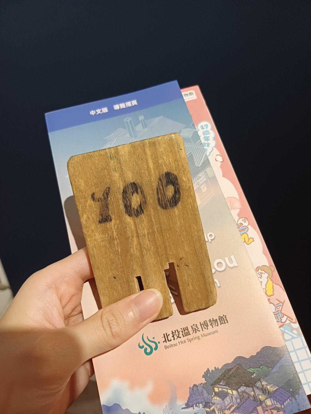 遊客手持北投溫泉博物館100號木製鞋栓,背景為導覽手冊