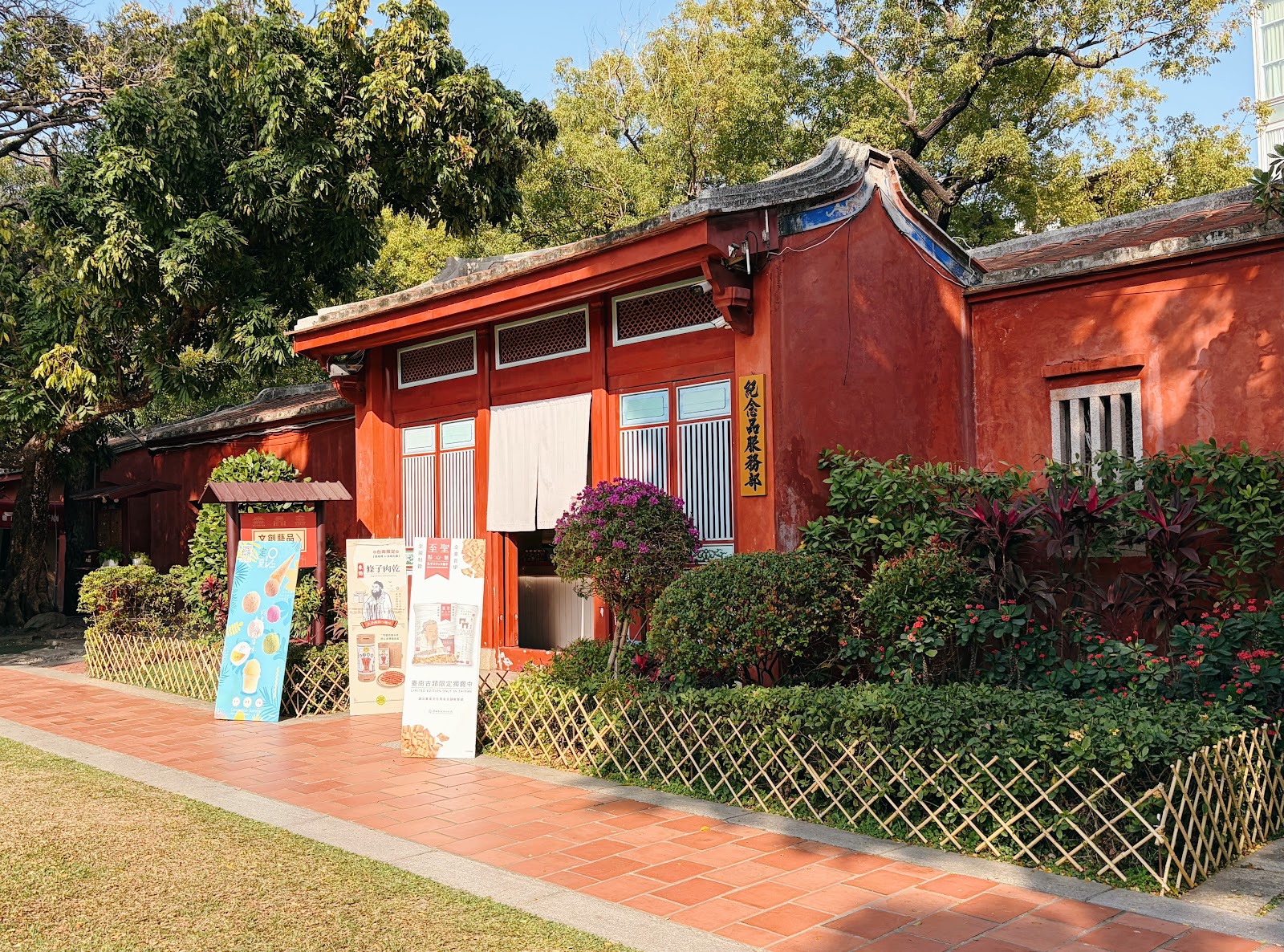 臺南孔廟側院紅色建築，門口紀念商品宣傳牌，花圃綠樹環繞