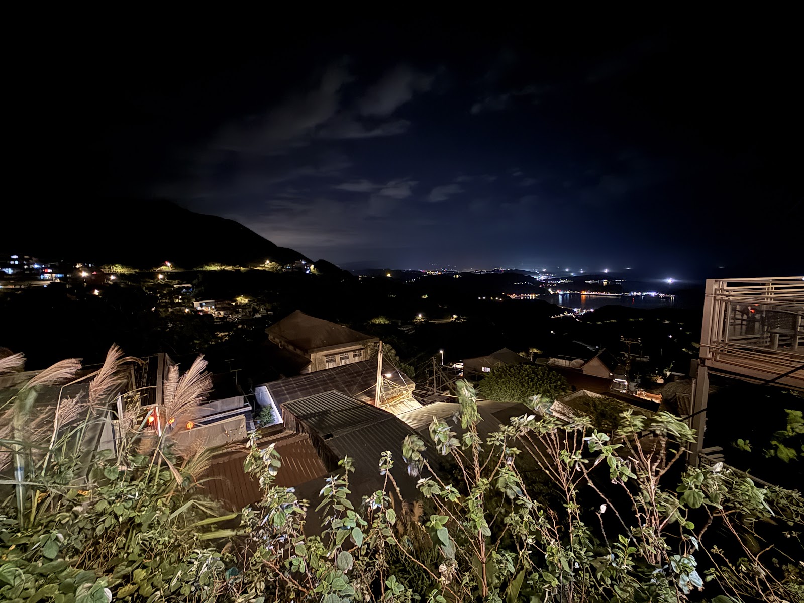 九份夜景廣角，前景屋頂與金黃芒草，遠處基隆港灣燈光閃爍
