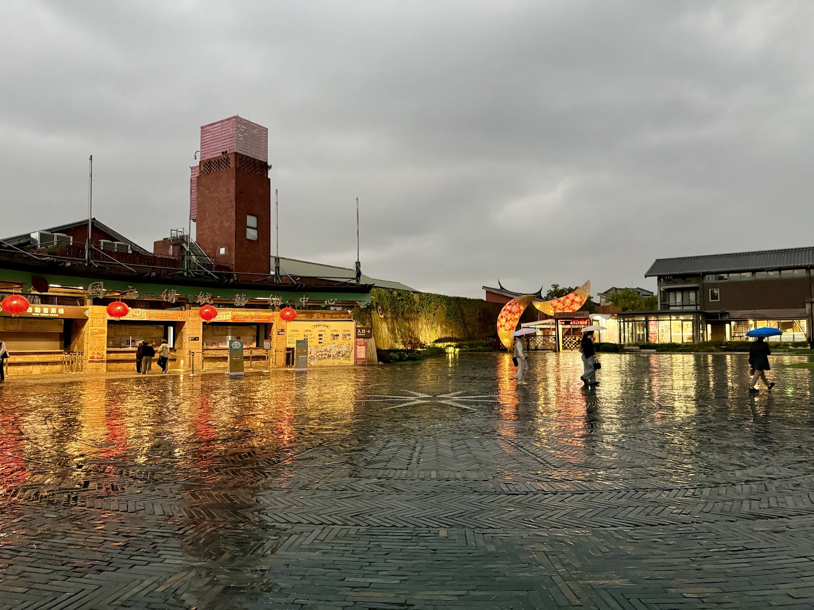 宜蘭傳藝園區廣場入口雨天景色，磚地積水倒映紅燈籠光芒，遊客撐傘遊覽