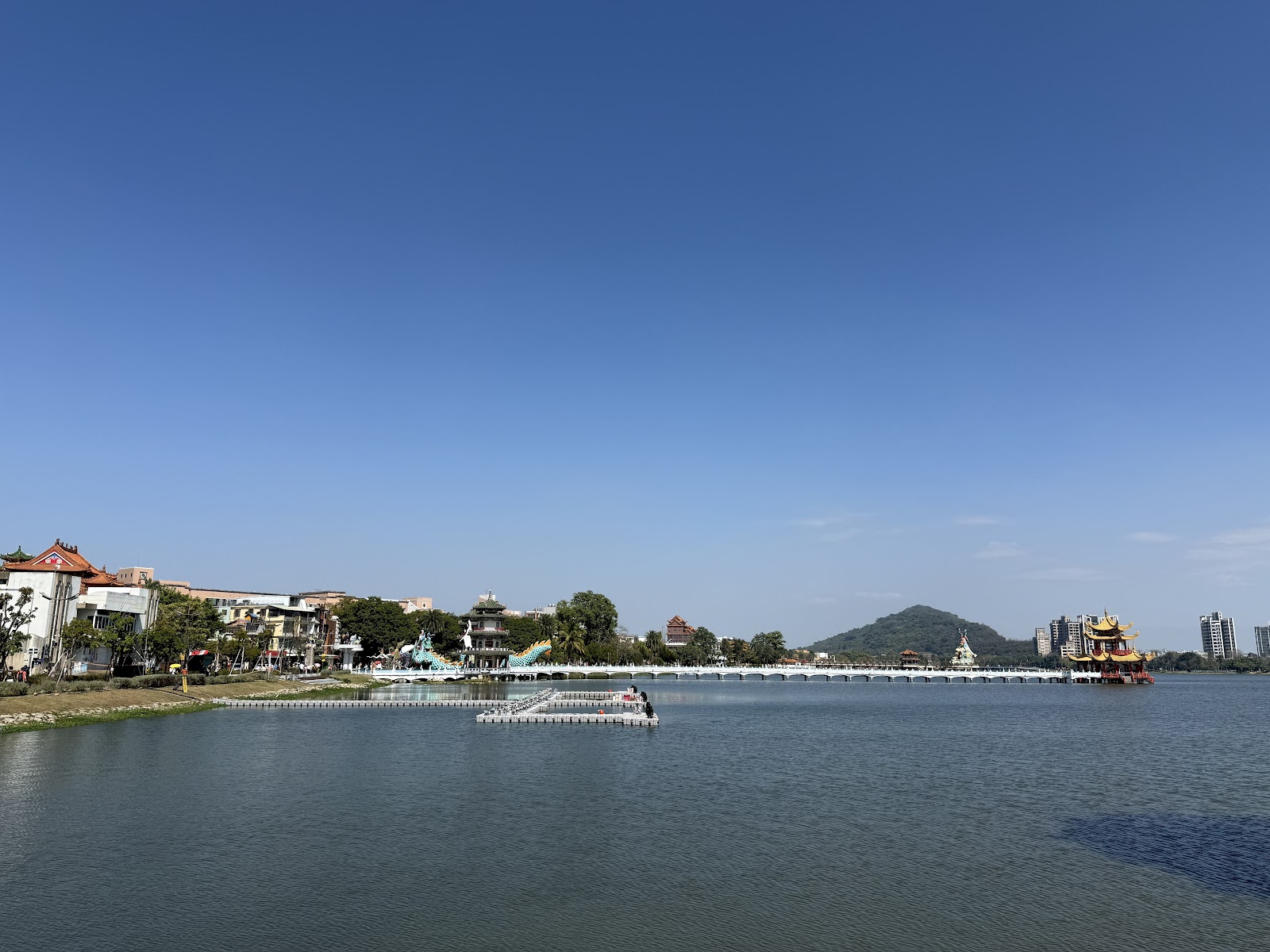 蓮池潭晴天湖景，棧道延伸湖中，廟宇與山丘遠景清晰