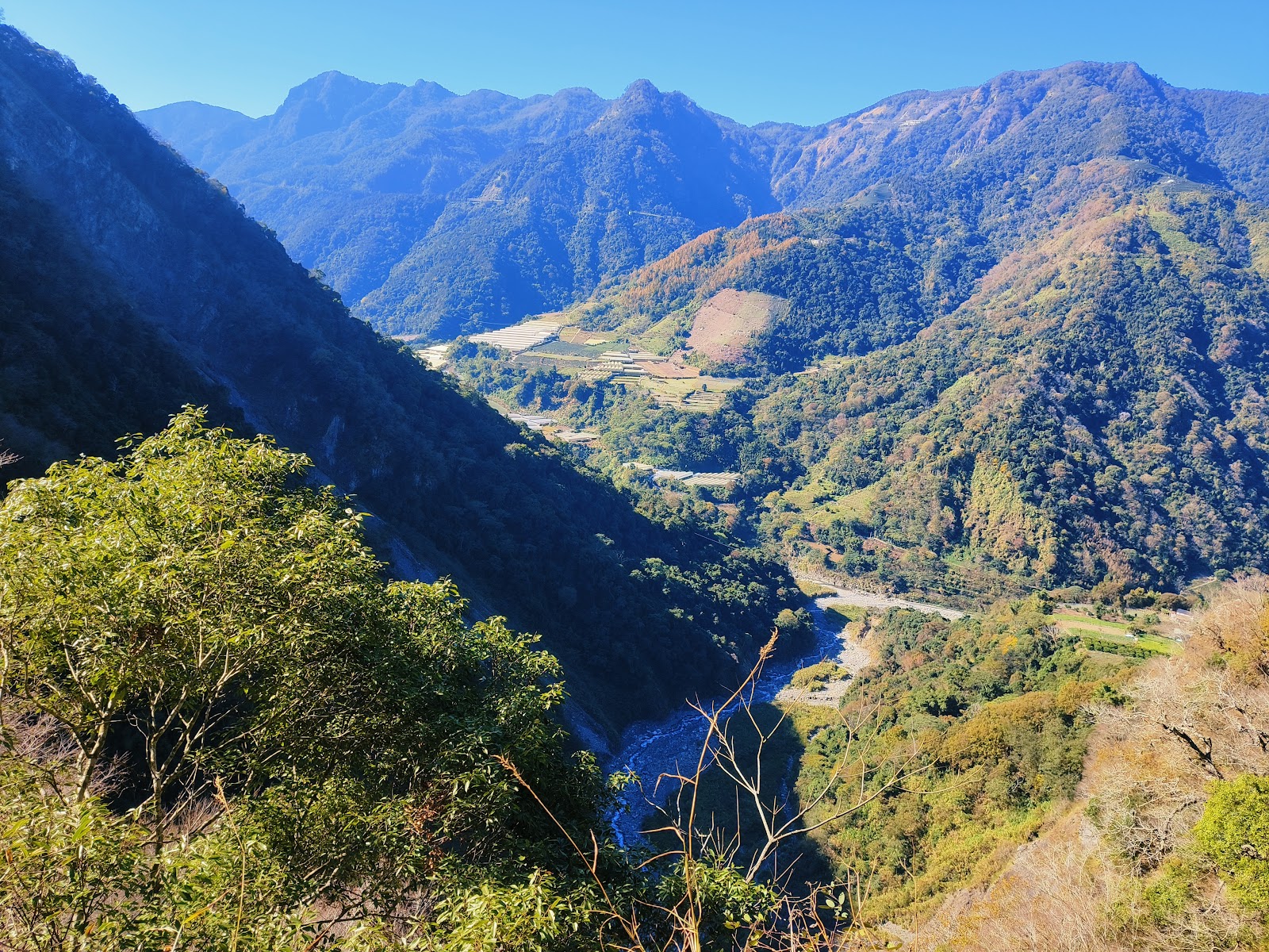 從高處俯瞰信義鄉山谷全景，溪流蜿蜒穿越山巒，秋色層疊，天空湛藍