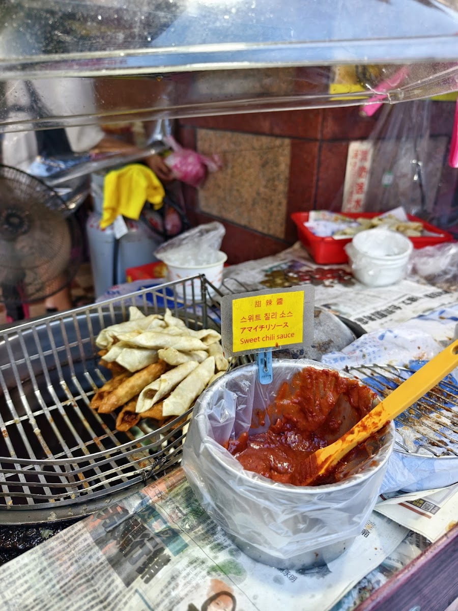 淡水老街路邊小吃攤，炸物放在烤架上，旁邊有甜辣醬小碗，多國語言標示