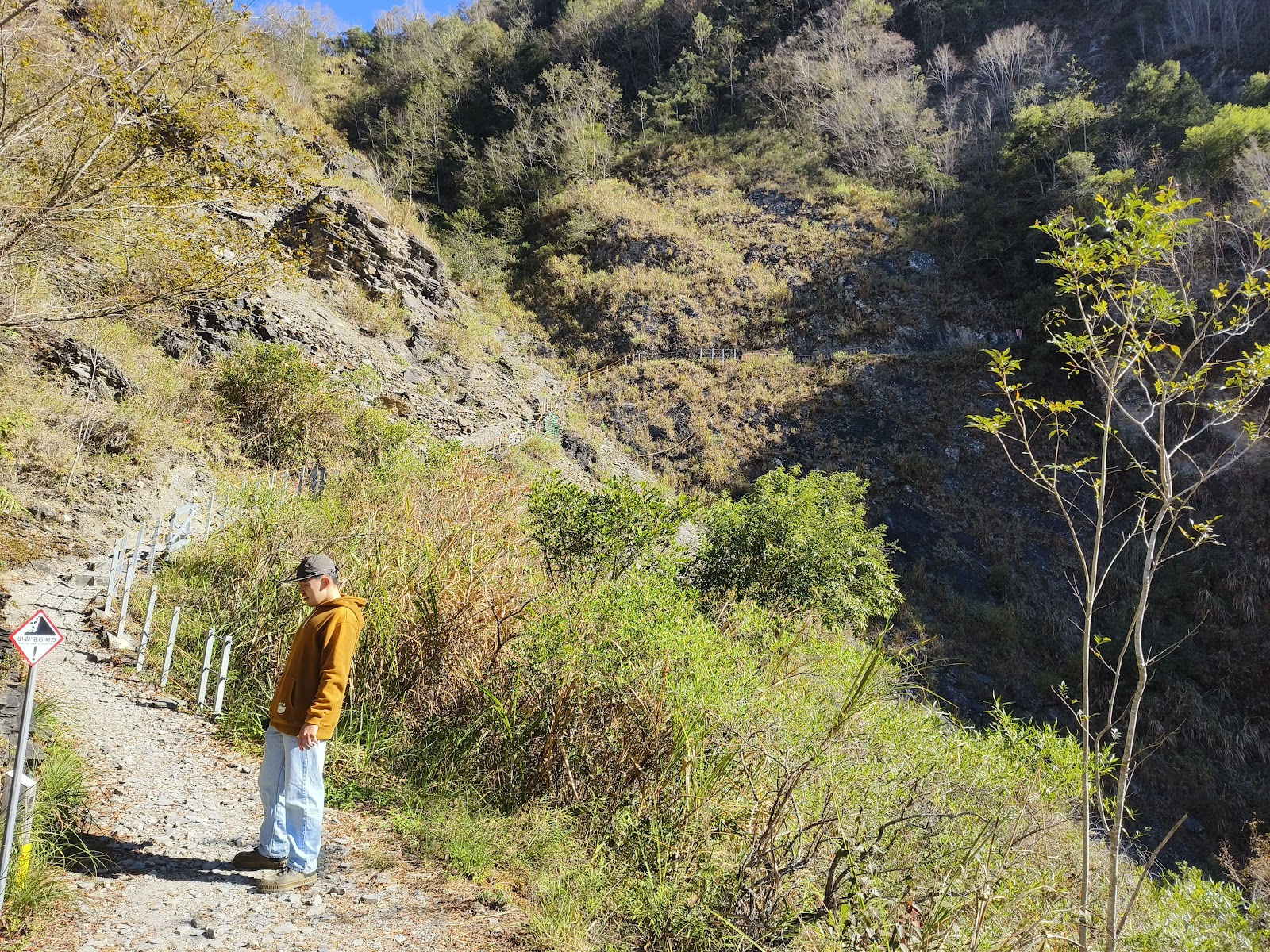 身穿橘色上衣的遊客站在山路旁俯瞰山谷，背景是乾燥的山坡地景