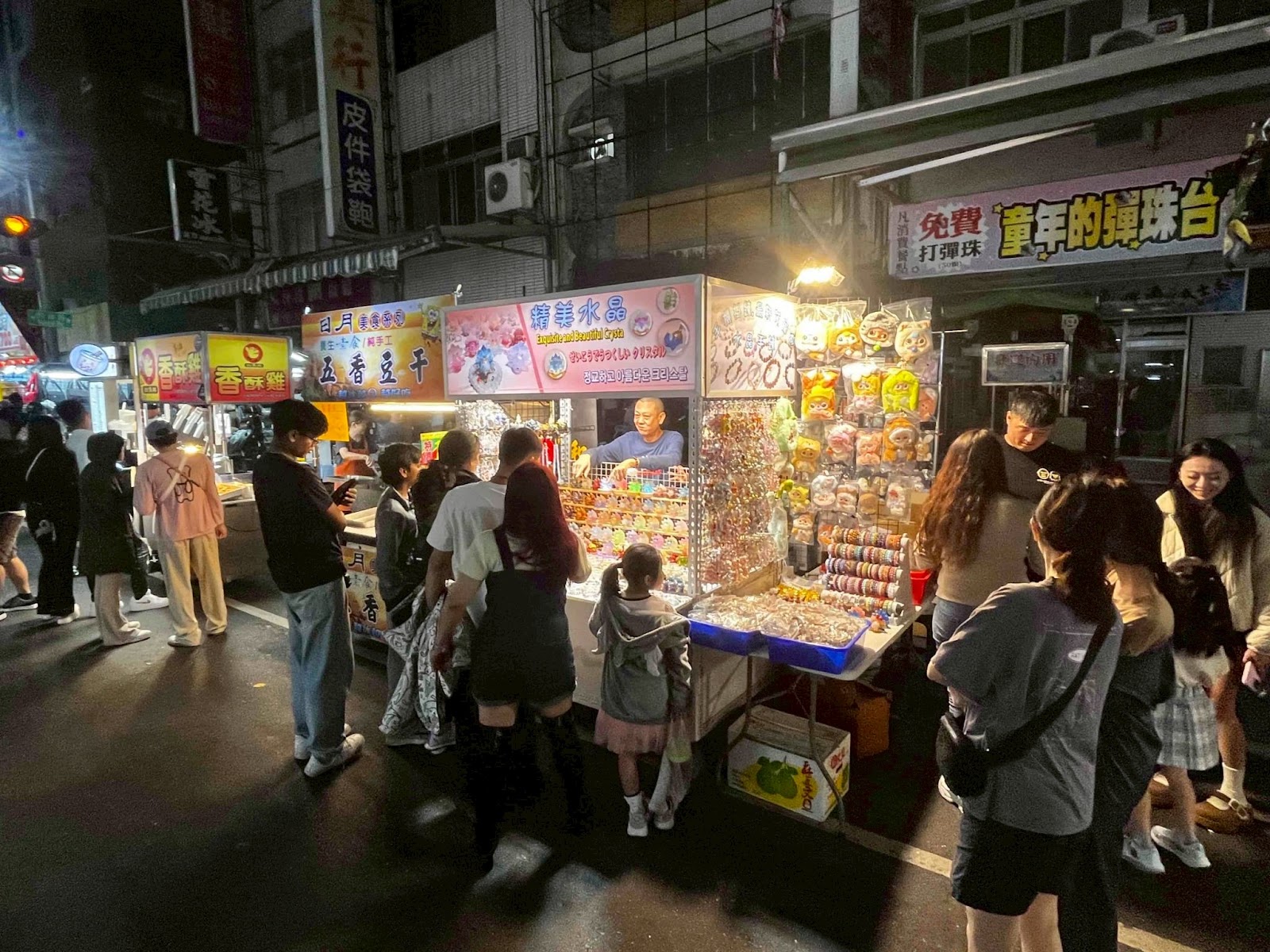夜市遊客聚集在糖果飾品攤前選購,旁邊有懷舊彈珠台