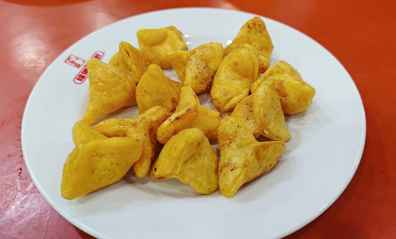 這張圖片中有一盤金黃色的炸餛飩，擺放在白色餐盤上。餛飩外皮香脆，旁邊是紅色的桌面，看起來十分可口。