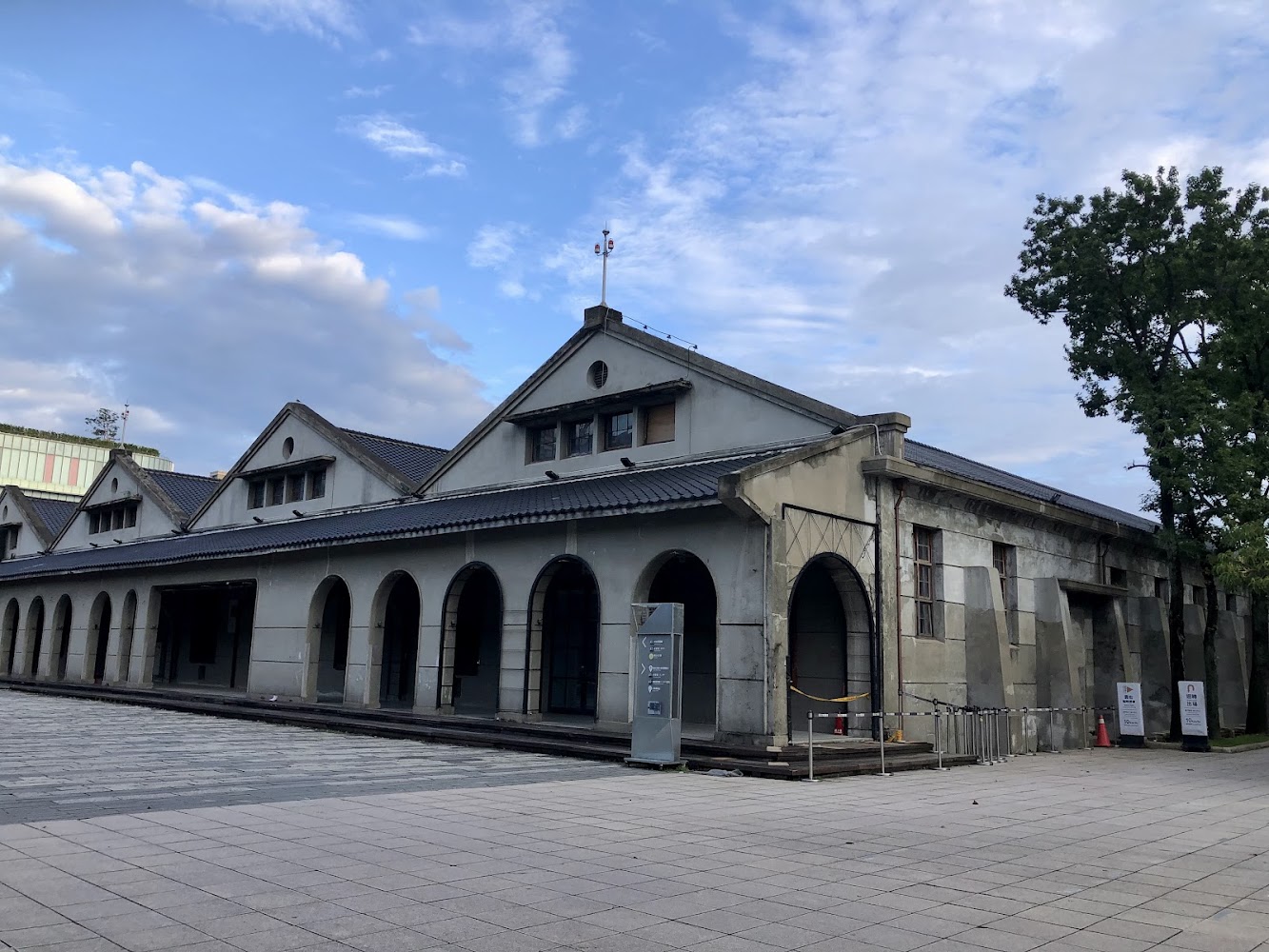 舊式倉庫建築，外觀有多個拱形門窗，牆面略顯斑駁，旁邊有樹木與廣場，天氣晴朗，整體氛圍安靜悠閒。