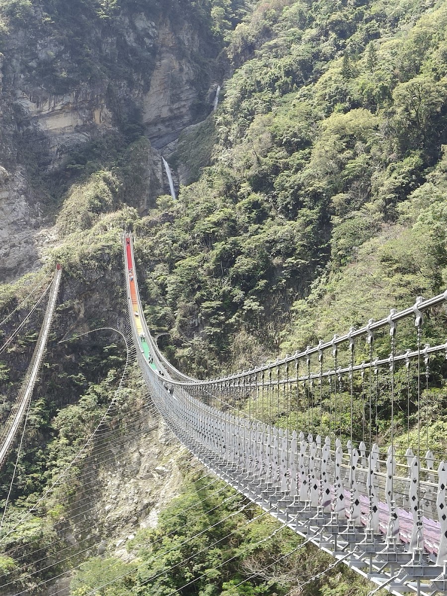 雙龍七彩吊橋側面全景，七彩橋面在深山峽谷中弧形延伸，背景可見瀑布飛瀑