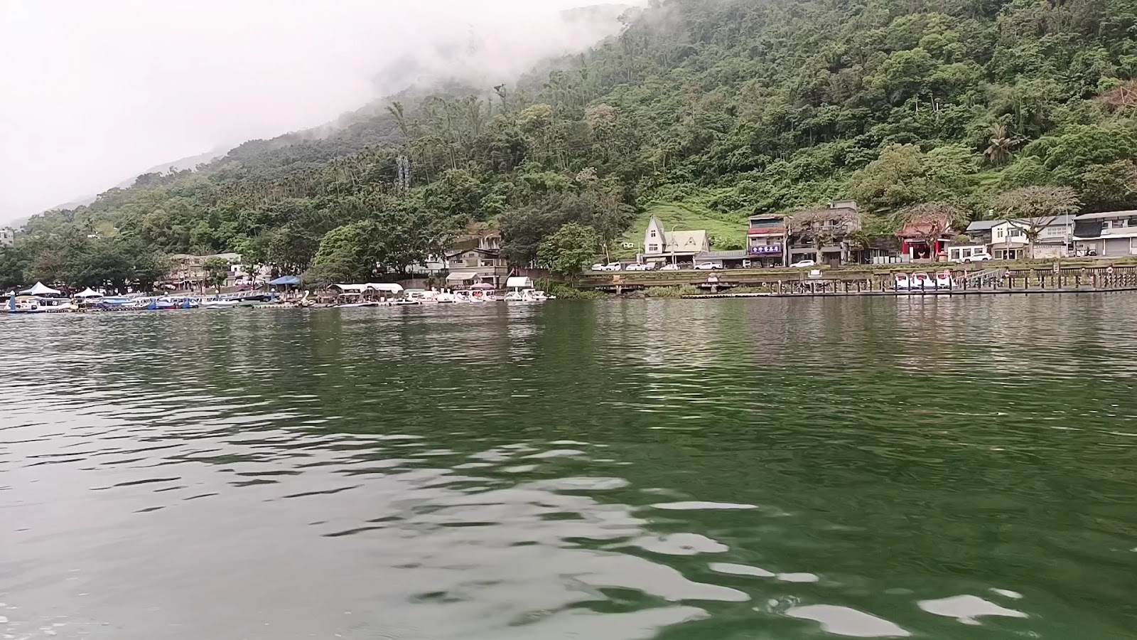 從水上視角拍攝鯉魚潭碼頭區，翠綠湖水前景，霧氣繚繞青山，對岸商店依山而建