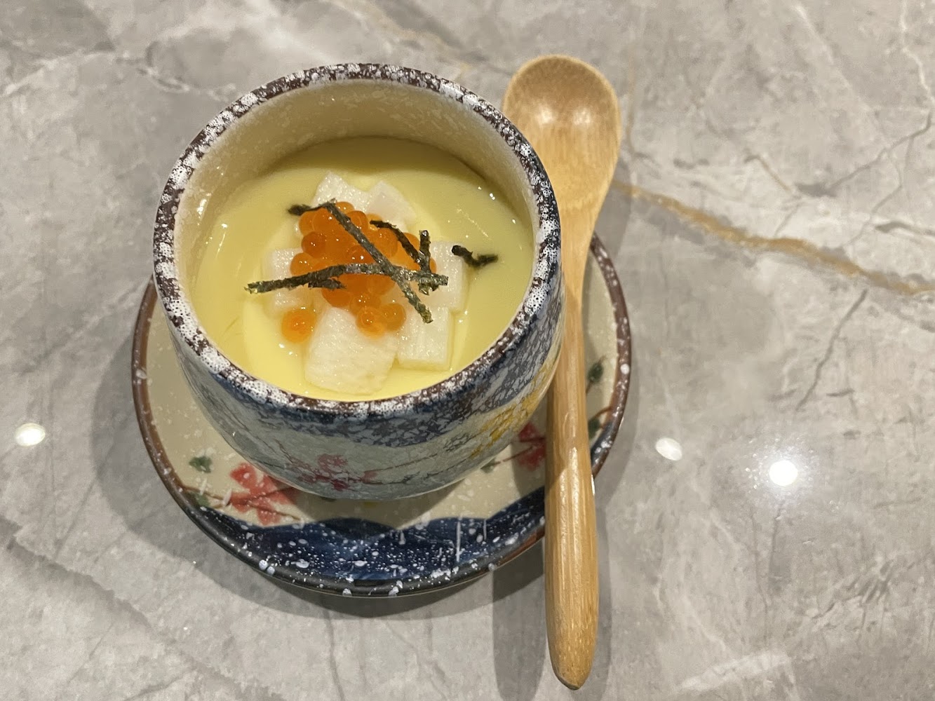 這張圖片主要是一杯日式茶碗蒸，表面有魚卵、白色食材和海苔絲點綴，旁邊放著木勺，擺放在花紋小碟上，看起來十分精緻。
