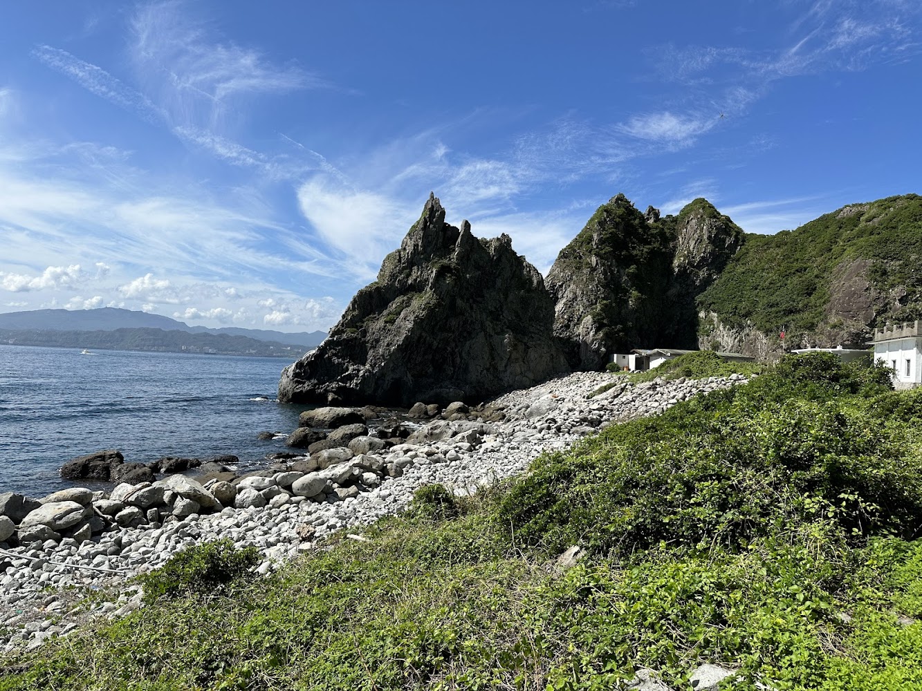 這張圖片呈現藍天白雲下的海岸景色，有奇特岩石、礫石灘和茂密綠意，還有幾間臨海小屋，整體自然風光壯麗。