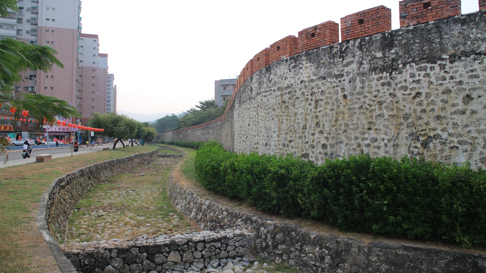 高雄左營舊城城牆側面長景,城牆延伸向遠方,前景護城河遺跡與綠色灌木,兩旁有現代住宅與燈籠