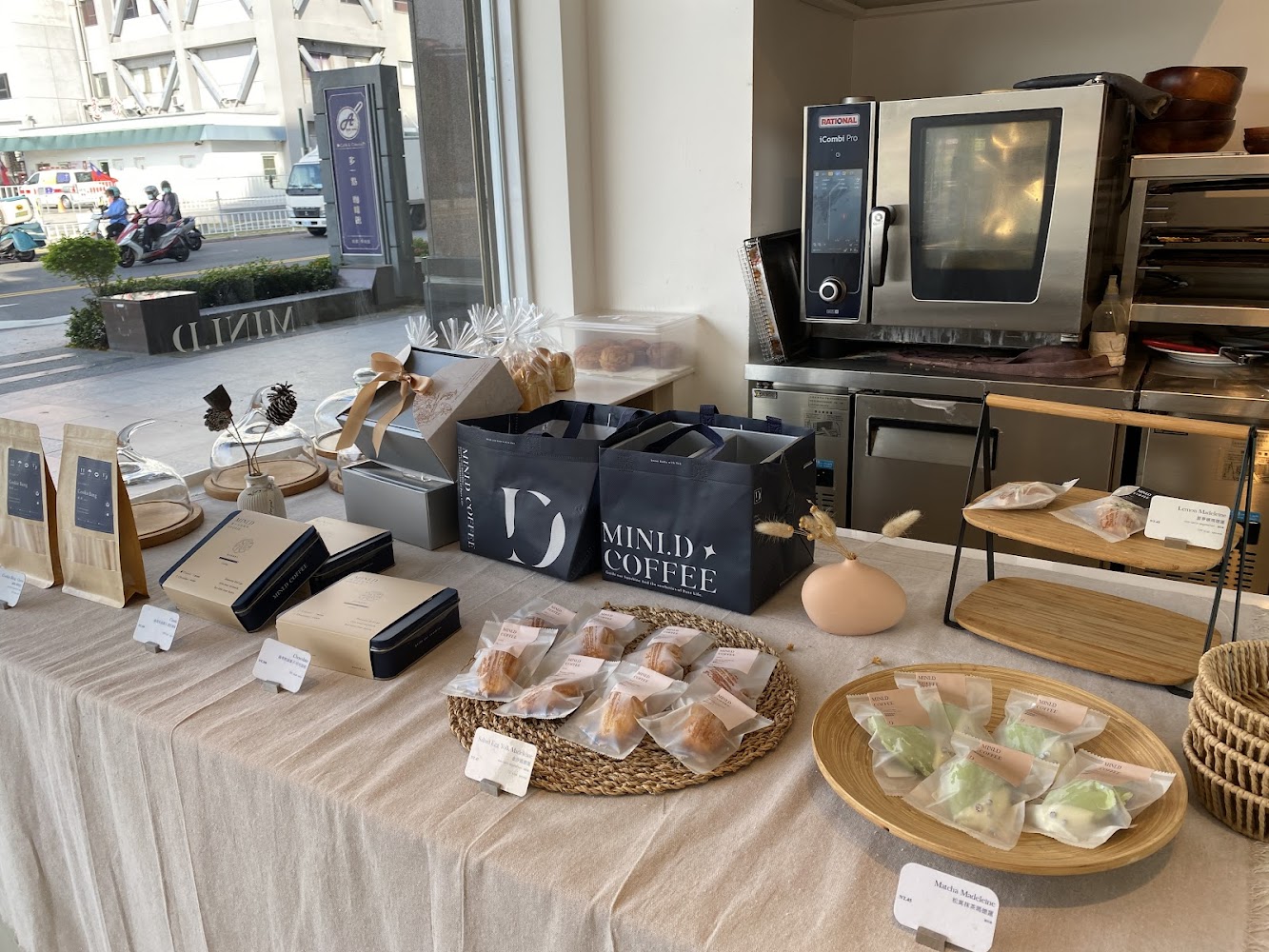 這張圖片展示了一家咖啡店的甜點陳列桌,上面有精緻包裝的蛋糕、馬德蓮、禮盒、咖啡豆包與手沖器具,整體擺設整齊有質感,後方有烤箱與烘焙設備。