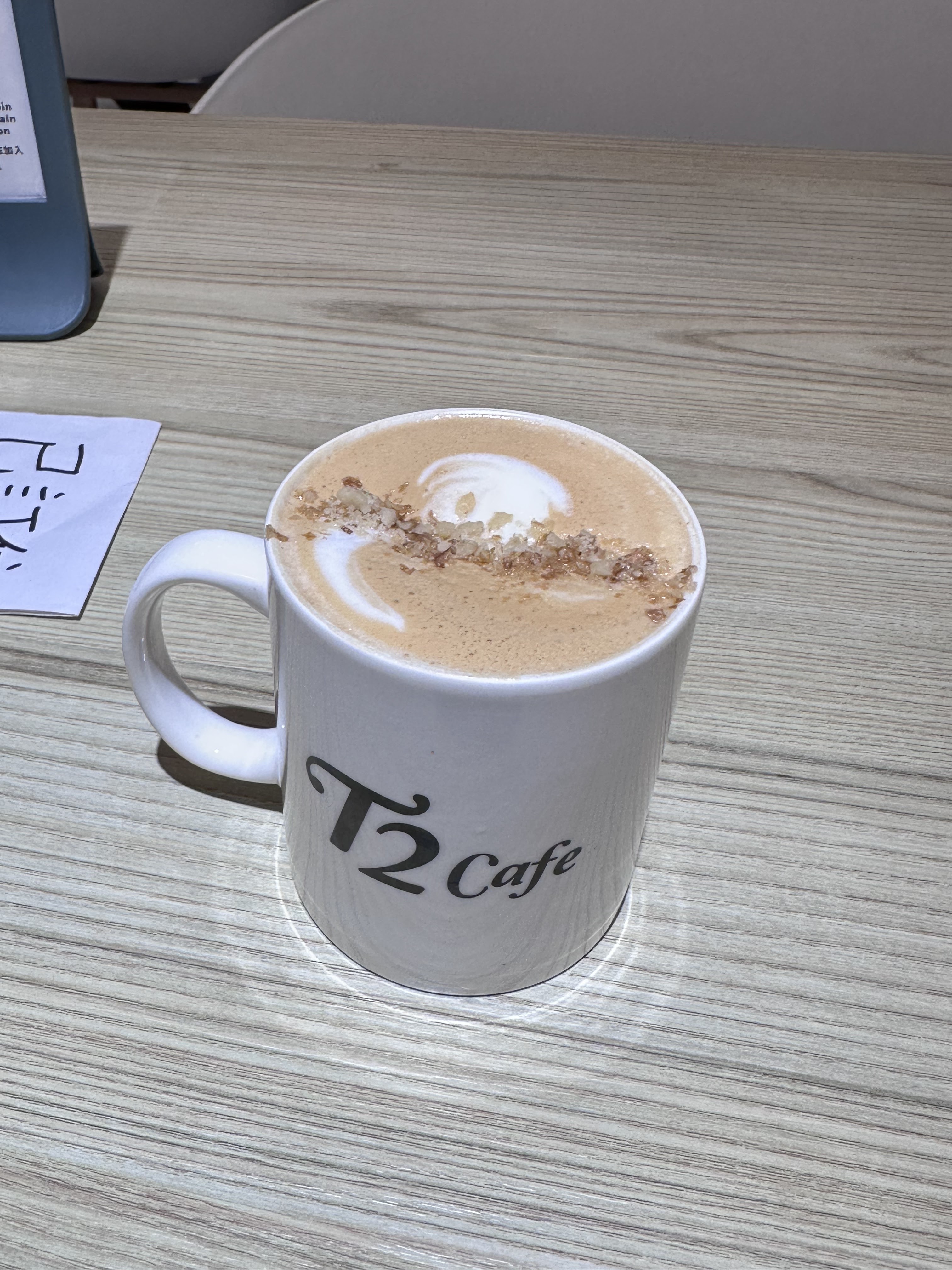 一杯拉花裝飾的熱飲，放在印有T2 Cafe字樣的白馬克杯裡