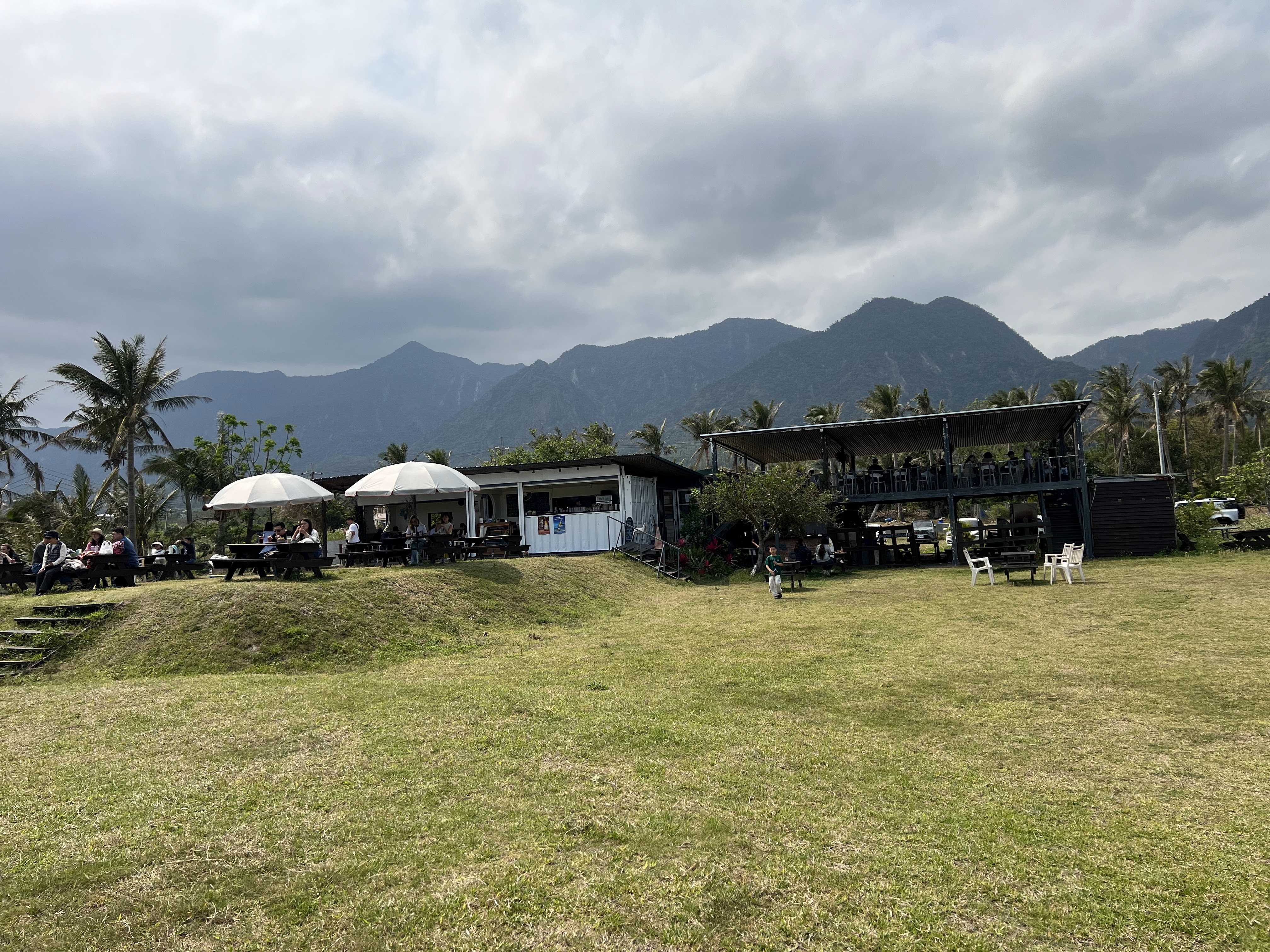 戶外草地上有桌椅與露天餐飲區，背景有山脈與棕櫚樹