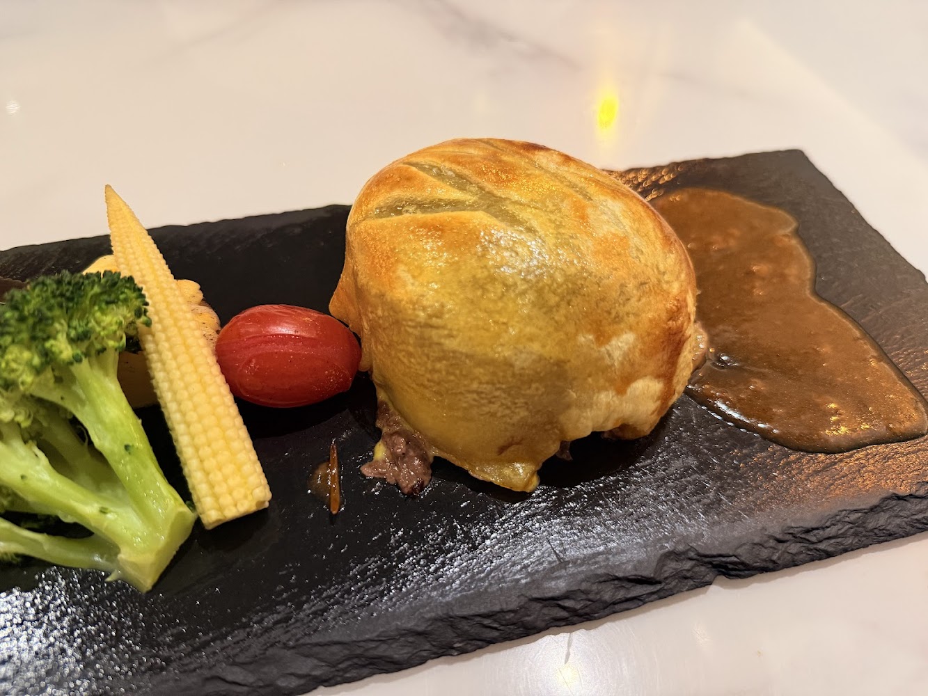 這張圖片主要呈現一道西餐料理，有金黃的酥皮包裹肉餡，搭配花椰菜、玉米筍及小番茄，旁邊有醬汁點綴。