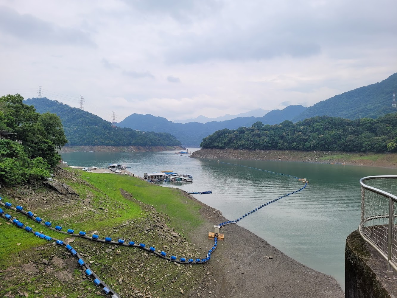 一處山區水庫，有寬廣的湖面和山林環繞。岸邊有浮標設施、幾艘船和碼頭。