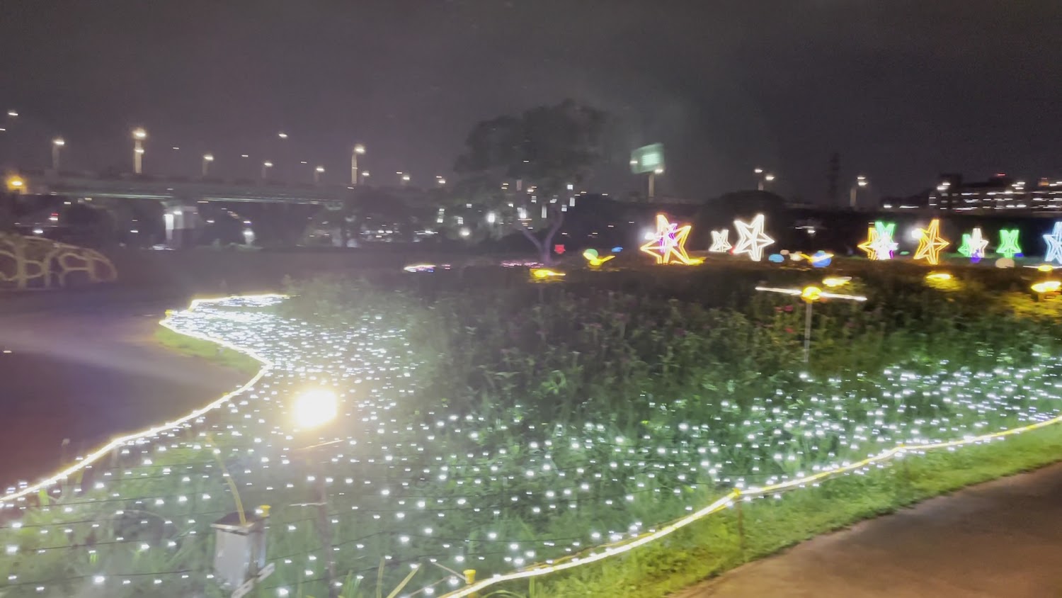 這張圖片展示夜間公園景色，地上和周圍被白色燈飾點綴，背景有多個五彩繽紛的星星造型燈飾，營造出浪漫且夢幻的氛圍。