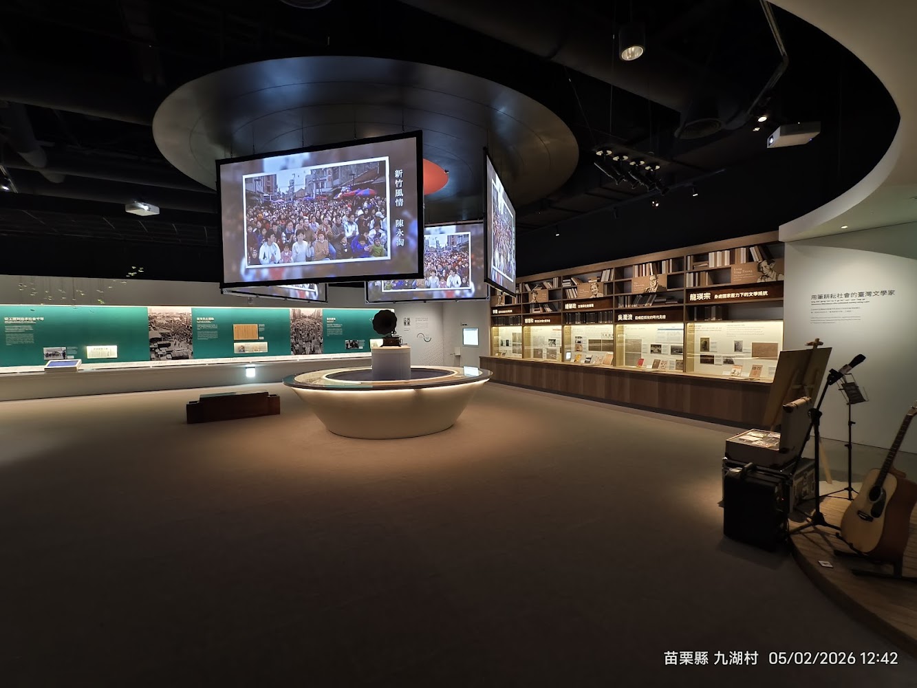 這張圖片展示了一間現代化展覽館，中央有大型懸掛螢幕播放影像，四周陳列著歷史文獻、展品和書籍，右下角擺放有吉他與音響設備，環境整潔且具科技感。