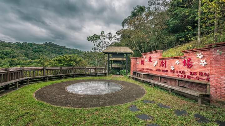 戶外露天劇場，前方有圓形表演區與紅磚牆座椅，背景山林環繞