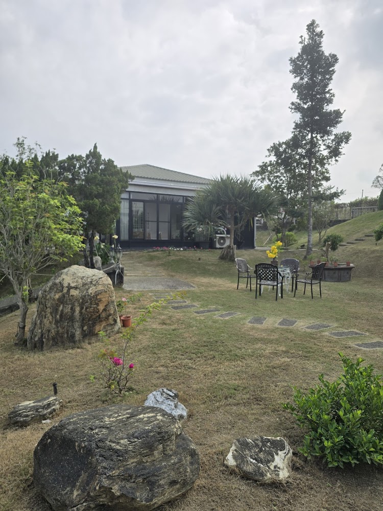 這張圖片主要展現了一個庭院，有大石頭、綠樹、草地及小花。中間擺放著桌椅，可供休憩，背景是一棟現代風格的房子，環境優美安靜。圖片沒有浮水印。