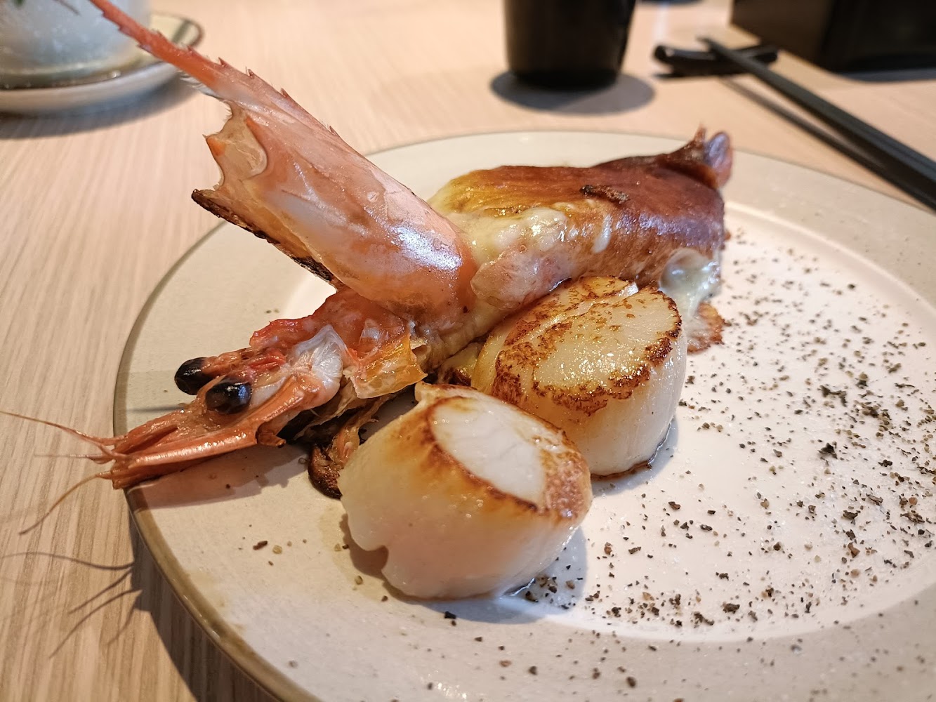 這張圖片主要呈現一道美食，包含一隻烤蝦和兩顆煎干貝，擺放在白色餐盤上，盤子邊緣灑有香草碎。