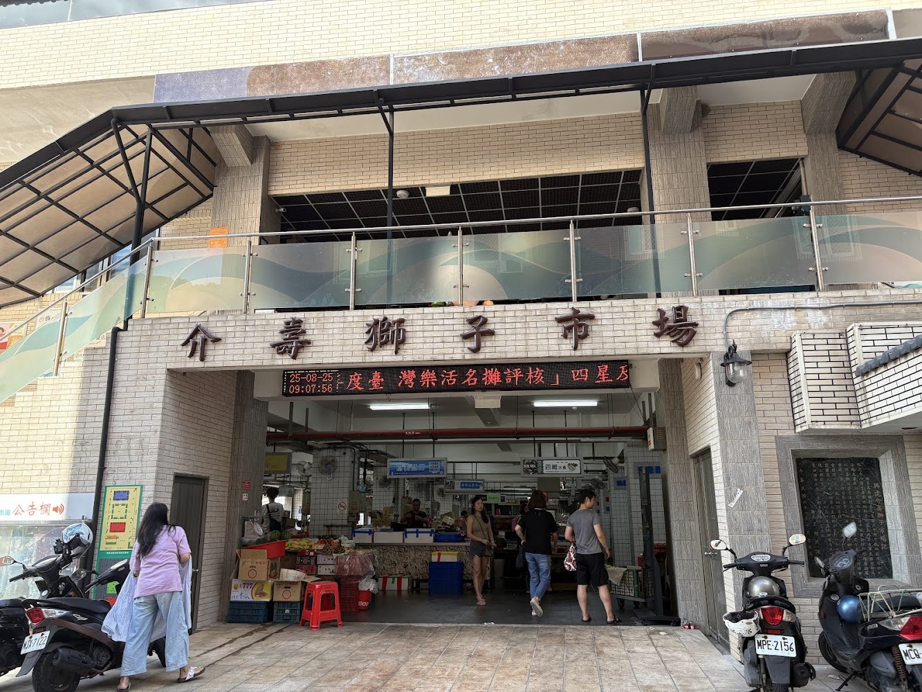 這是一間名為『獅子市場』的傳統市場，入口有攤販、顧客及停放的機車，市場內部可見賣菜與日常雜貨的攤位，是社區民眾購物的重要據點。