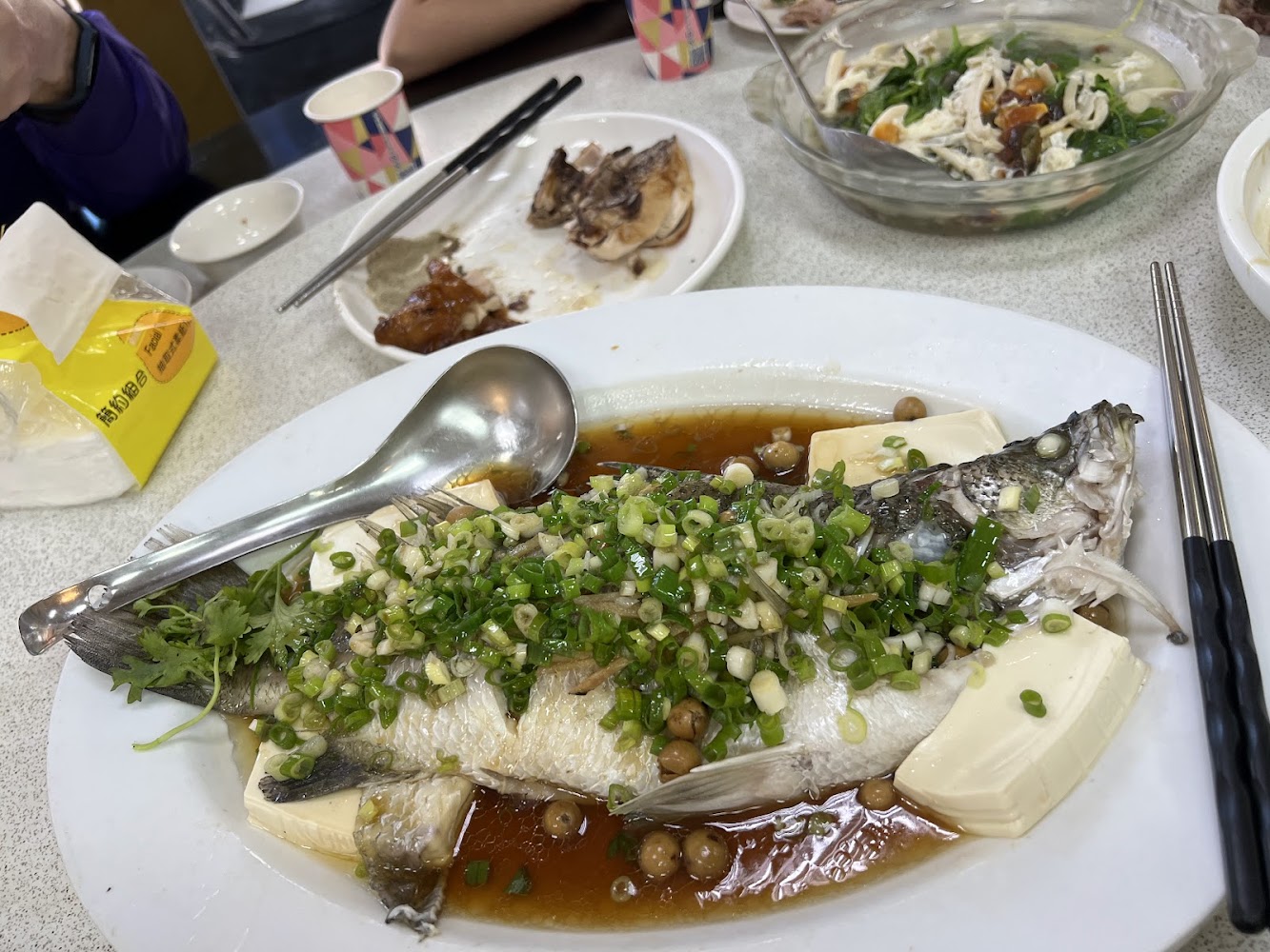 一盤清蒸魚，魚上鋪有大量蔥蒜與醬汁，旁邊搭配豆腐