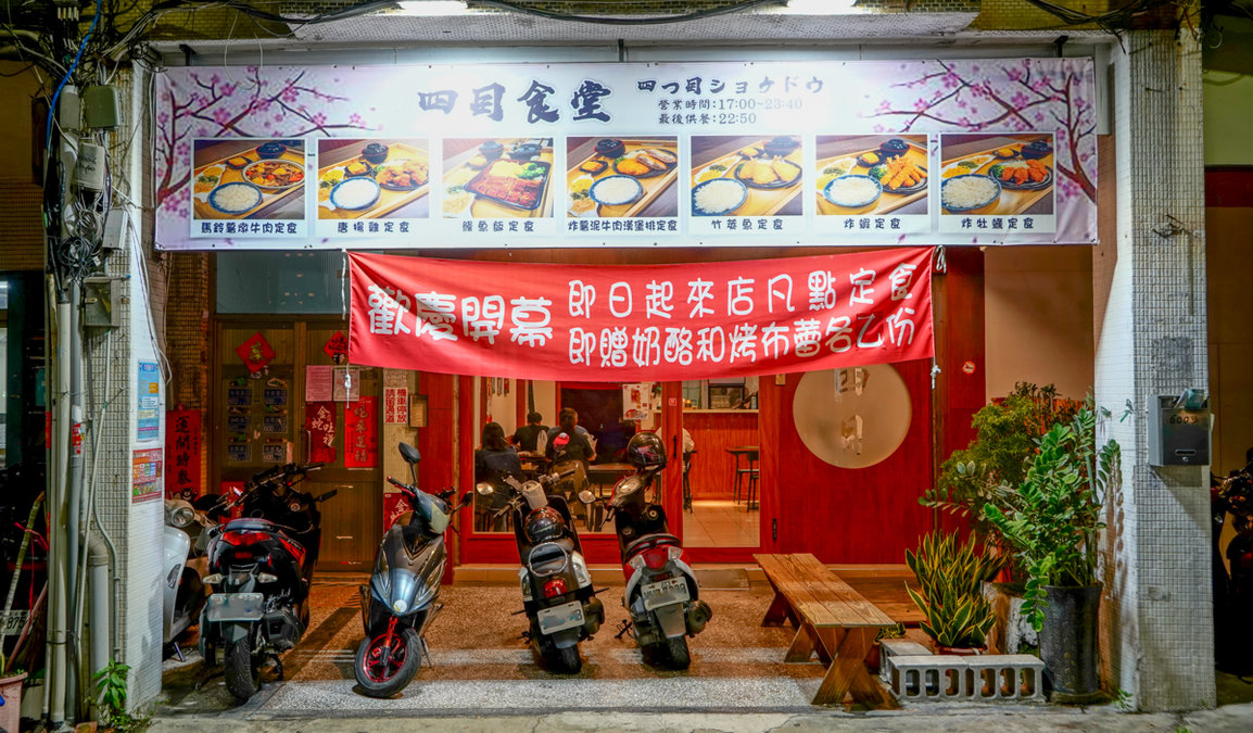 這是一間位於街頭的日式定食餐廳門口，外頭停放著數台機車。店面招牌上展示多種定食圖片，並懸掛紅色開幕布條，宣傳點定食可贈送奶酪和布蕾。