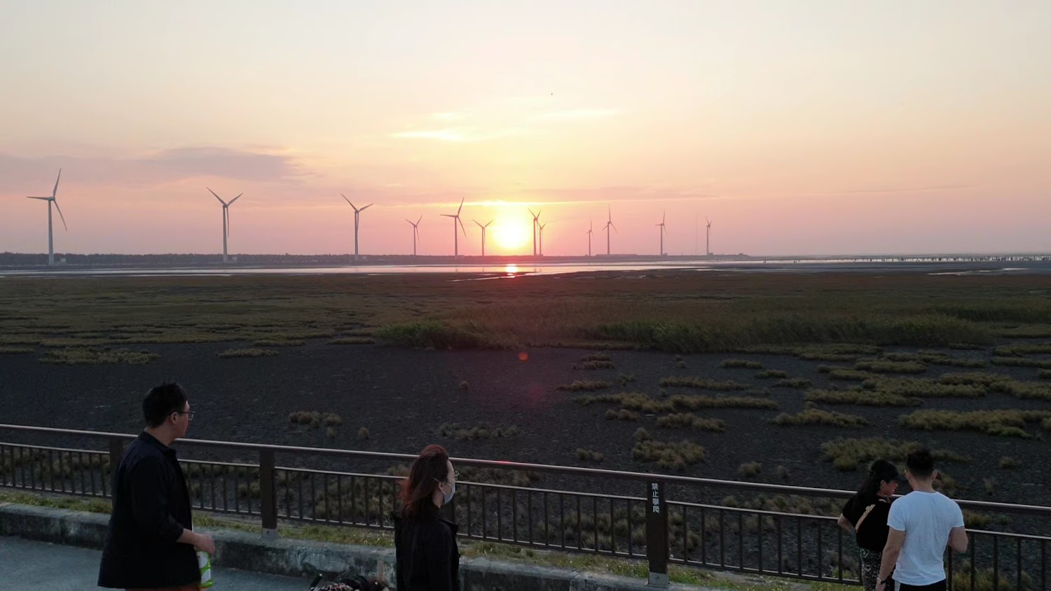 夕陽下的濕地前景有欄杆邊的遊客,遠景是風力發電機