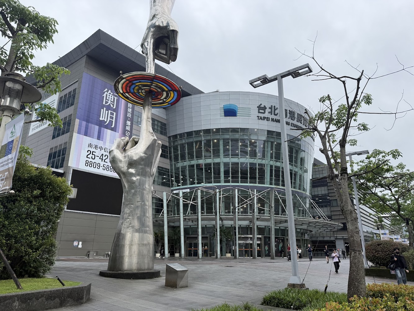 台北南港展覽館建築外觀，前景有金屬雕塑，雕塑為兩隻手托舉著彩色圓盤，四周環繞著綠樹與行人，環境現代且寬敞。