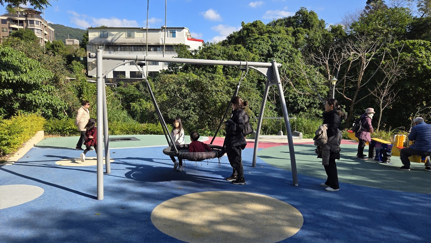 戶外遊樂場有鞦韆、蹺蹺板和多位大人與小孩正在遊玩